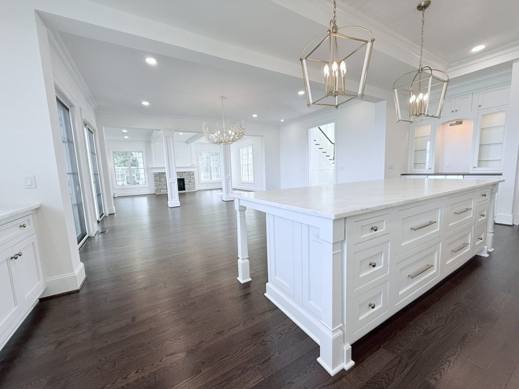 Kitchen island with white cabinetry and brass lantern pendant lights, opening into a great room with a stone fireplace
