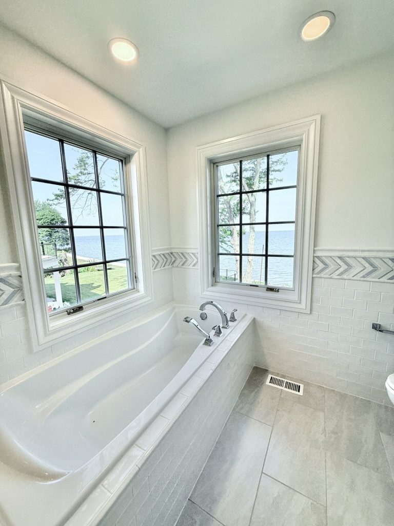 Freestanding soaking tub with wall-mount fixtures, dual windows with a neighborhood and lake view, and a herringbone tile accent band