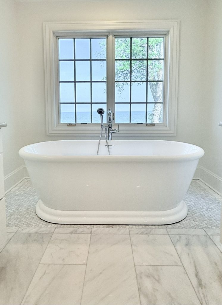 Freestanding soaking tub with a floor-mount tub filler beneath a large window overlooking the lake