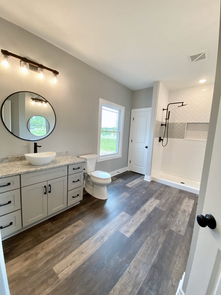 The Golden Primary Bathroom with Gray Vanity, Walk-In Shower, Window Light & Wood-Look Flooring