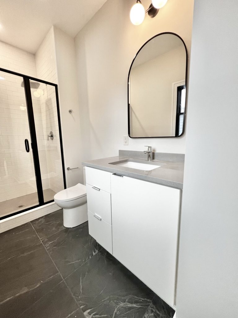 The Cedar White Vanity with Arched Mirror, Black-Framed Shower, Dark Tile Flooring & Chrome Faucet