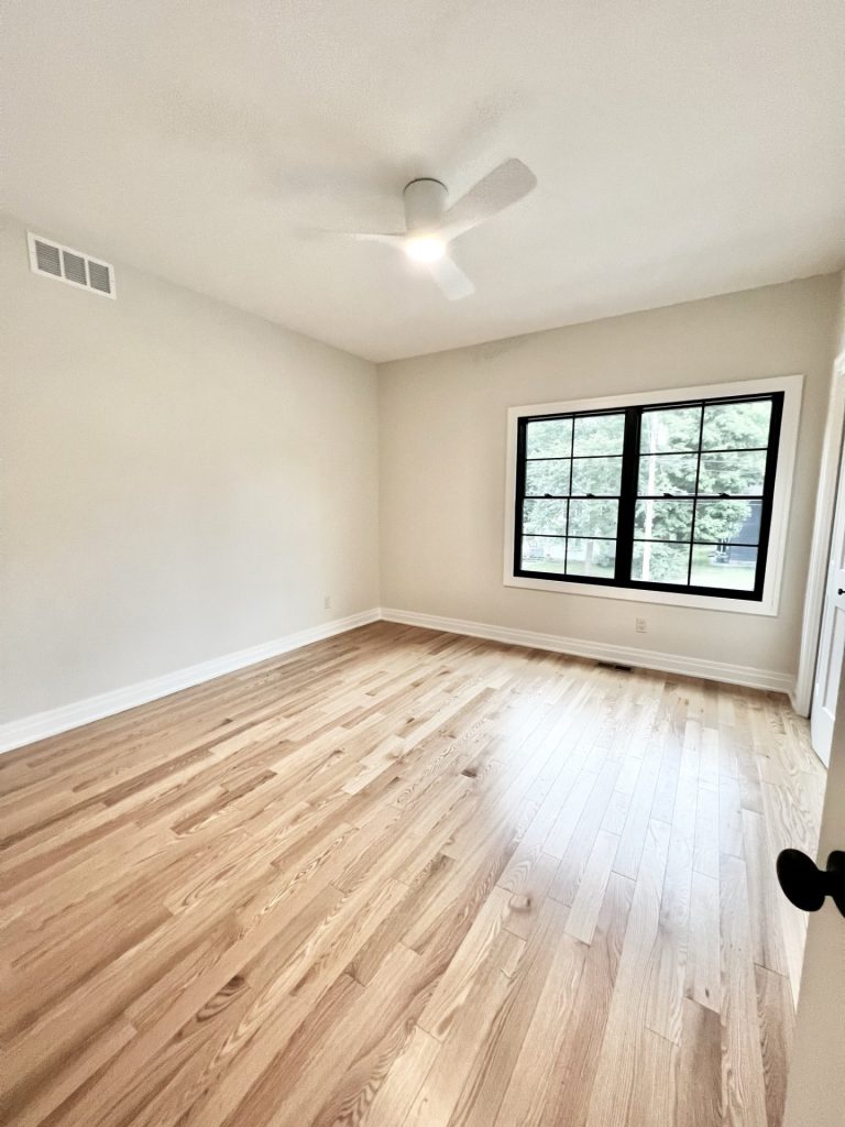 The Cedar Bedroom with Hardwood Flooring, Ceiling Fan, Large Black-Framed Window & Neutral Walls