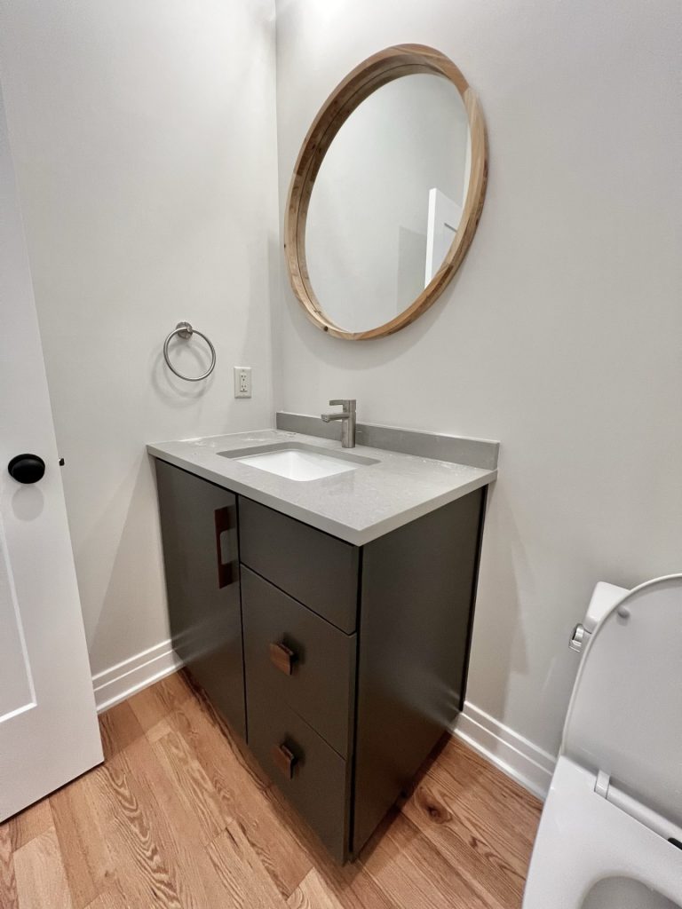 The Cedar Powder Room Vanity with Dark Cabinetry, Round Mirror, Quartz Countertop & Hardwood Flooring