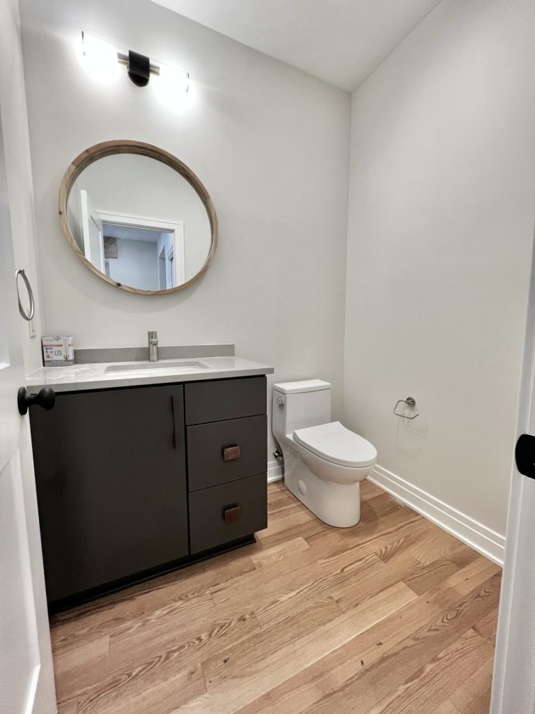 The Cedar Powder Room with Dark Vanity, Round Mirror, Quartz Countertop & Hardwood Flooring