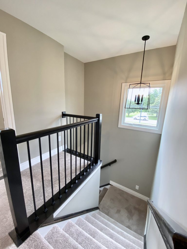 The Weston Carpeted Staircase View From the Landing with Dark Balusters, a Window and a Modern Chandelier