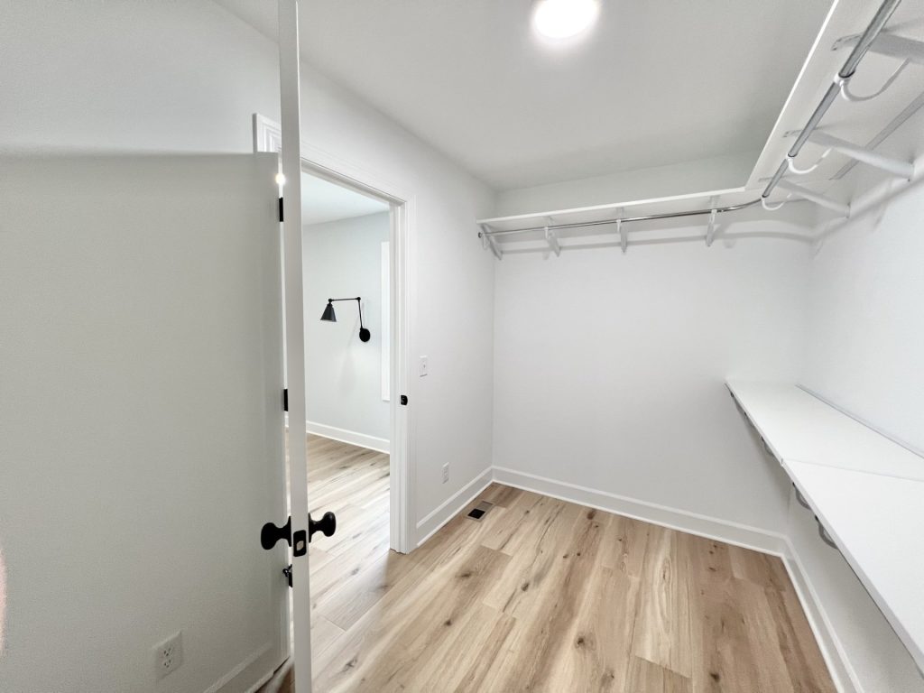 The Willow Walk-in Closet in Light Colors with Wrap-Around Shelving and Wood Flooring with Open Door to Master Bedroom