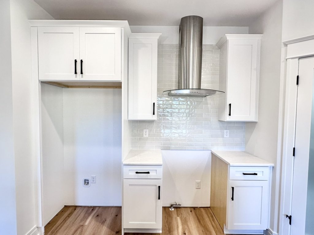 In-progress kitchen installation with white shaker cabinets and a stainless range hood over gray subway tile awaiting backsplash completion
