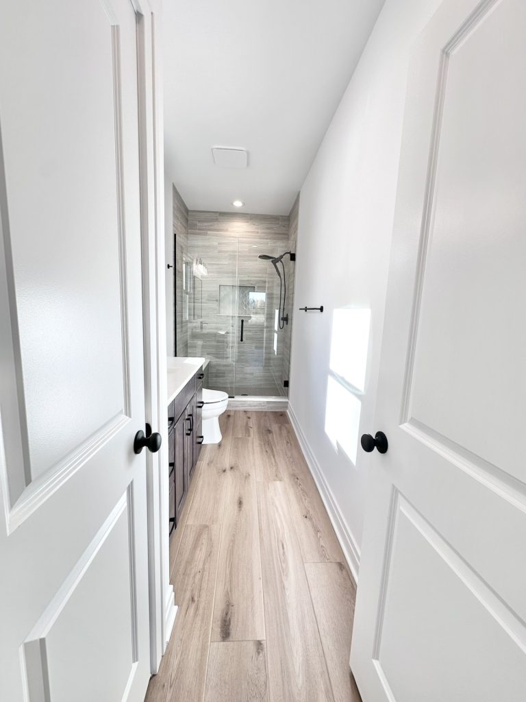 Hallway view into a primary bathroom with a walk-in tiled shower, dark vanity, and wood-look flooring