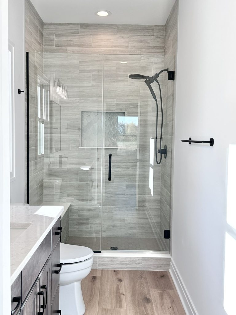 Walk-in tiled shower under construction with frameless glass door, gray plank tile walls, black fixtures, and a hexagon-tile niche