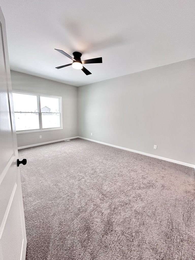 Empty bedroom with gray carpet, black ceiling fan, and a window overlooking a snowy subdivision