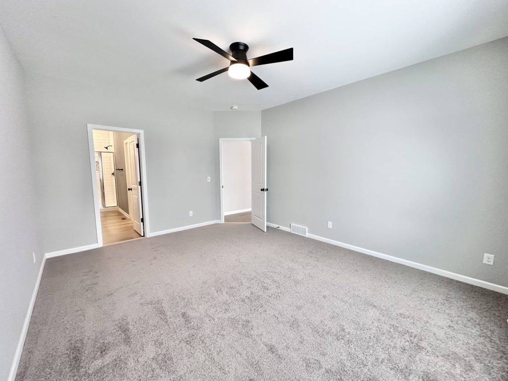 Empty bedroom with gray carpet, black ceiling fan, and doors leading to an en-suite bathroom and hallway